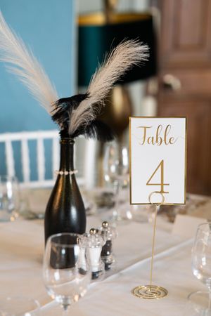 Elegant wedding table setting with gold table number 4 and black bottle decorated with white feathers as centerpiece.