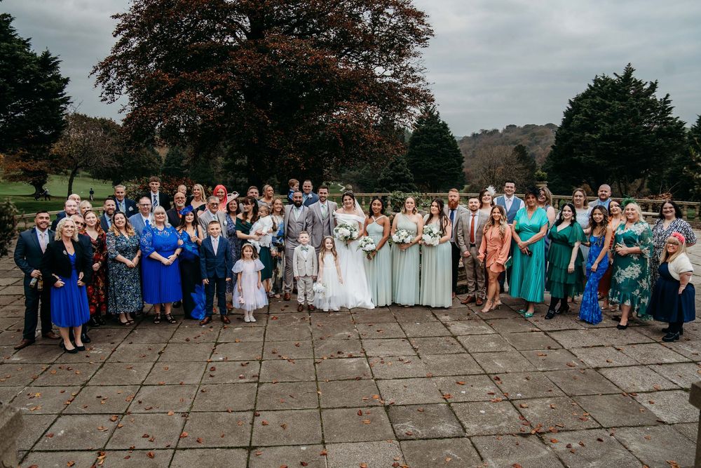 Group gathered for outdoor wedding photo on stone patio under cloudy sky with autumn trees in background.