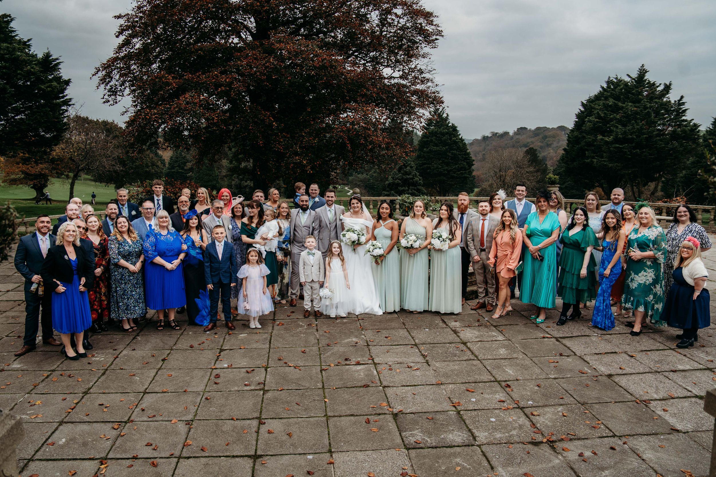 Group gathered for outdoor wedding photo on stone patio under cloudy sky with autumn trees in background.