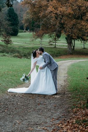 A romantic wedding photo captures a loving embrace on a tree-lined path in an autumn park with fall foliage.