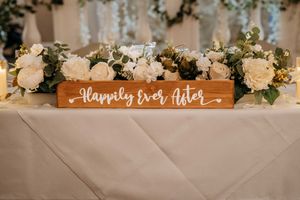 Wooden wedding sign reading 'Happily Ever After' decorated with white roses and greenery on a light tablecloth.