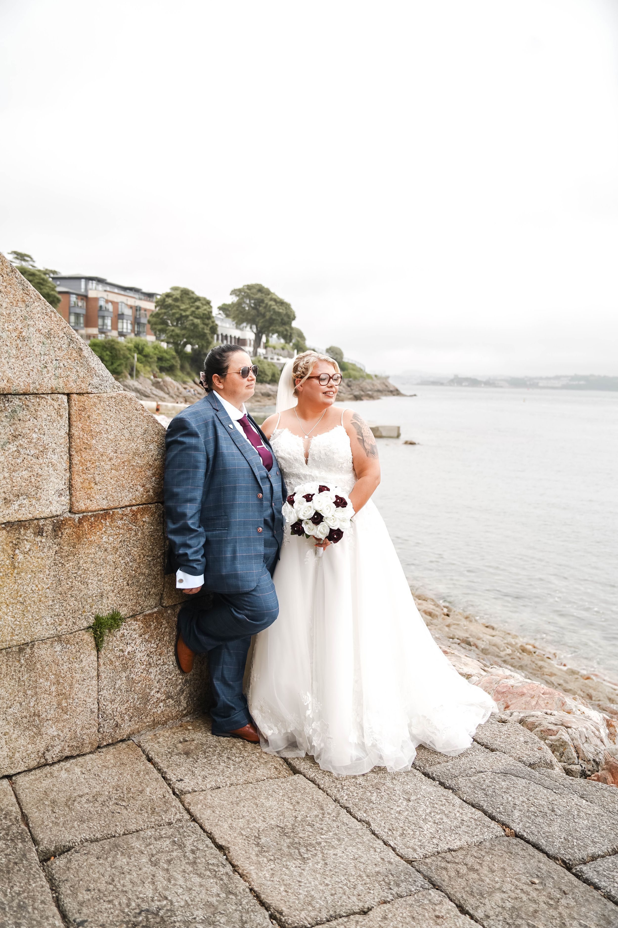 A couple in wedding attire sharing intimate moments along a stone seawall with ocean views in the background.