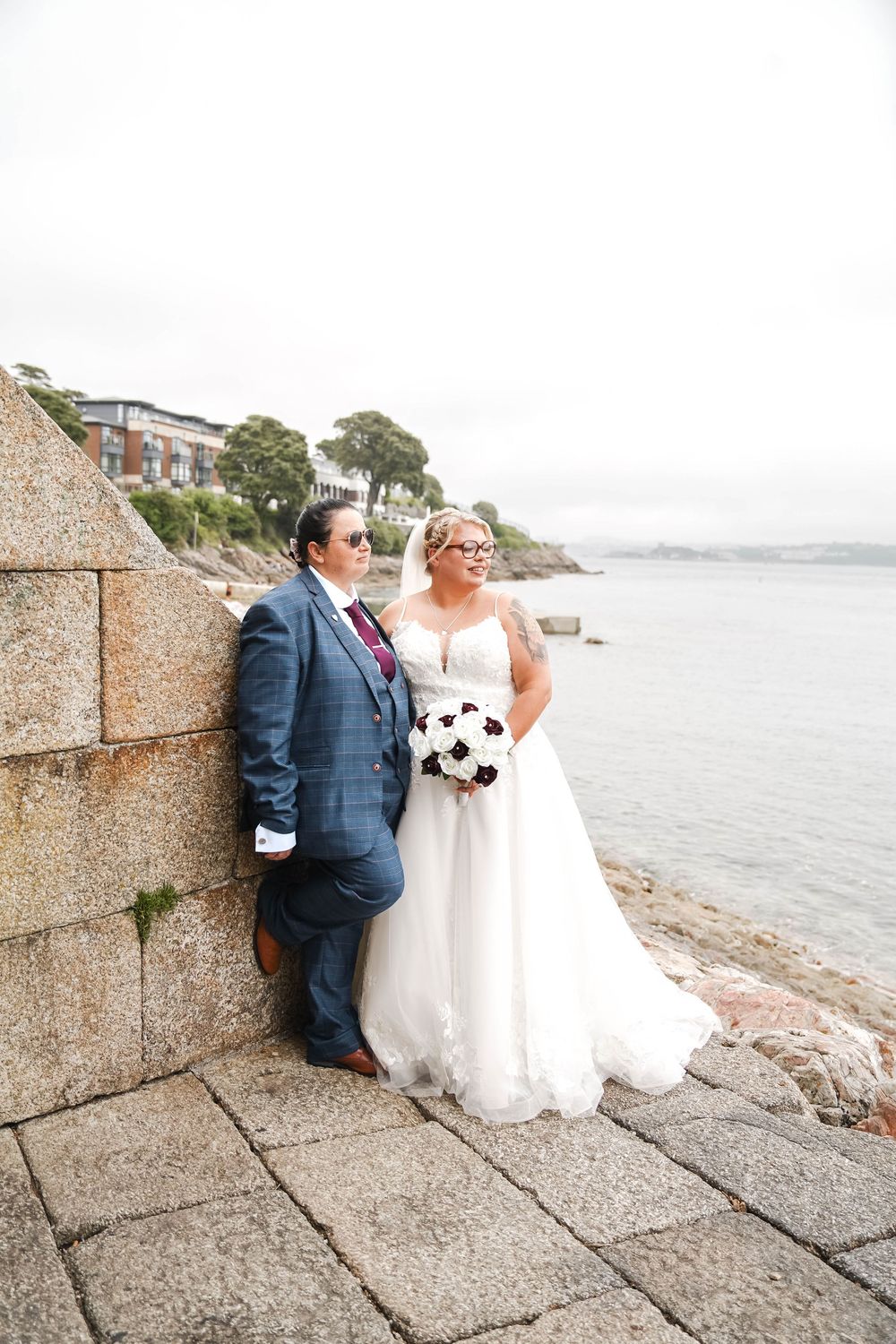 A couple in wedding attire sharing intimate moments along a stone seawall with ocean views in the background.