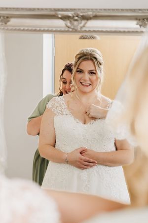 A bride wearing a beautiful lace wedding dress and headpiece smiles warmly during her wedding preparation.