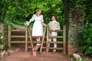 A couple in wedding attire stands on opposite sides of a wooden fence gate in a lush green forest setting.