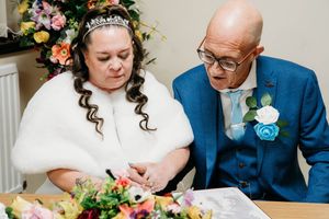 A bride in a white fur wrap and tiara sits at a table with an older gentleman in a blue suit during a wedding ceremony.