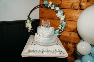 White tiered wedding cake with blue floral decorations and arch display against rustic wooden wall.