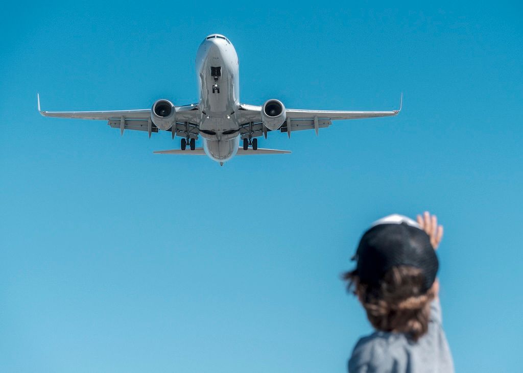 A boy is waving at a jumbo jet as it is landing and flying over his head.