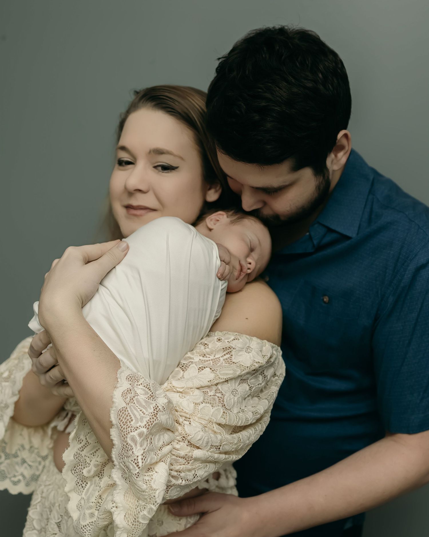 Parents tenderly cradling and kissing their sleeping newborn baby while dressed in blue and off-white lace outfits.
