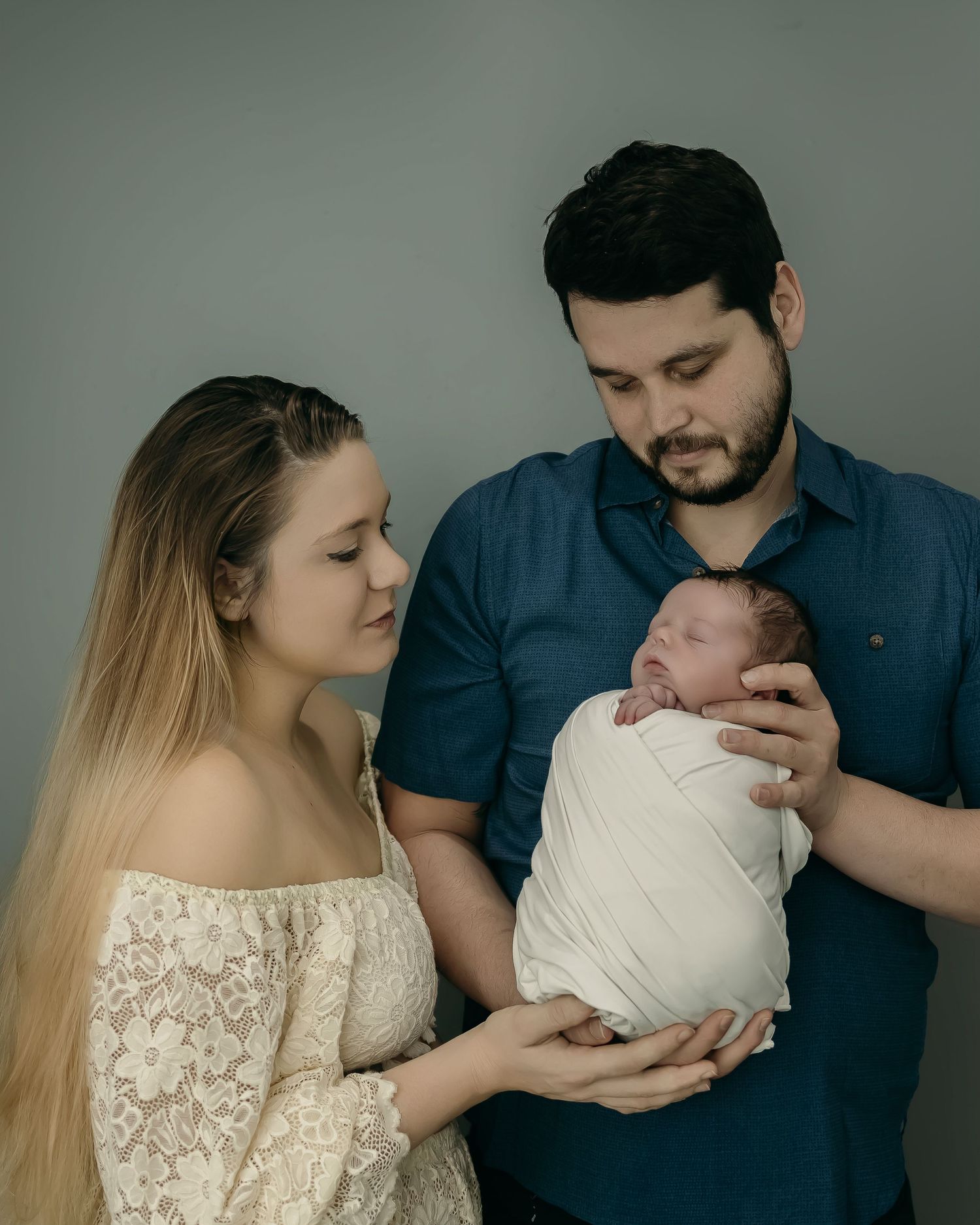 A couple cradles their newborn baby wrapped in white swaddling cloth against a grey background.