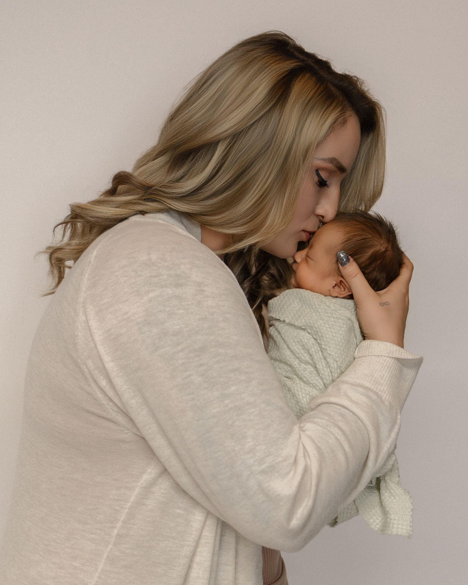 Woman in cream sweater tenderly kisses sleeping newborn baby wrapped in light mint blanket.