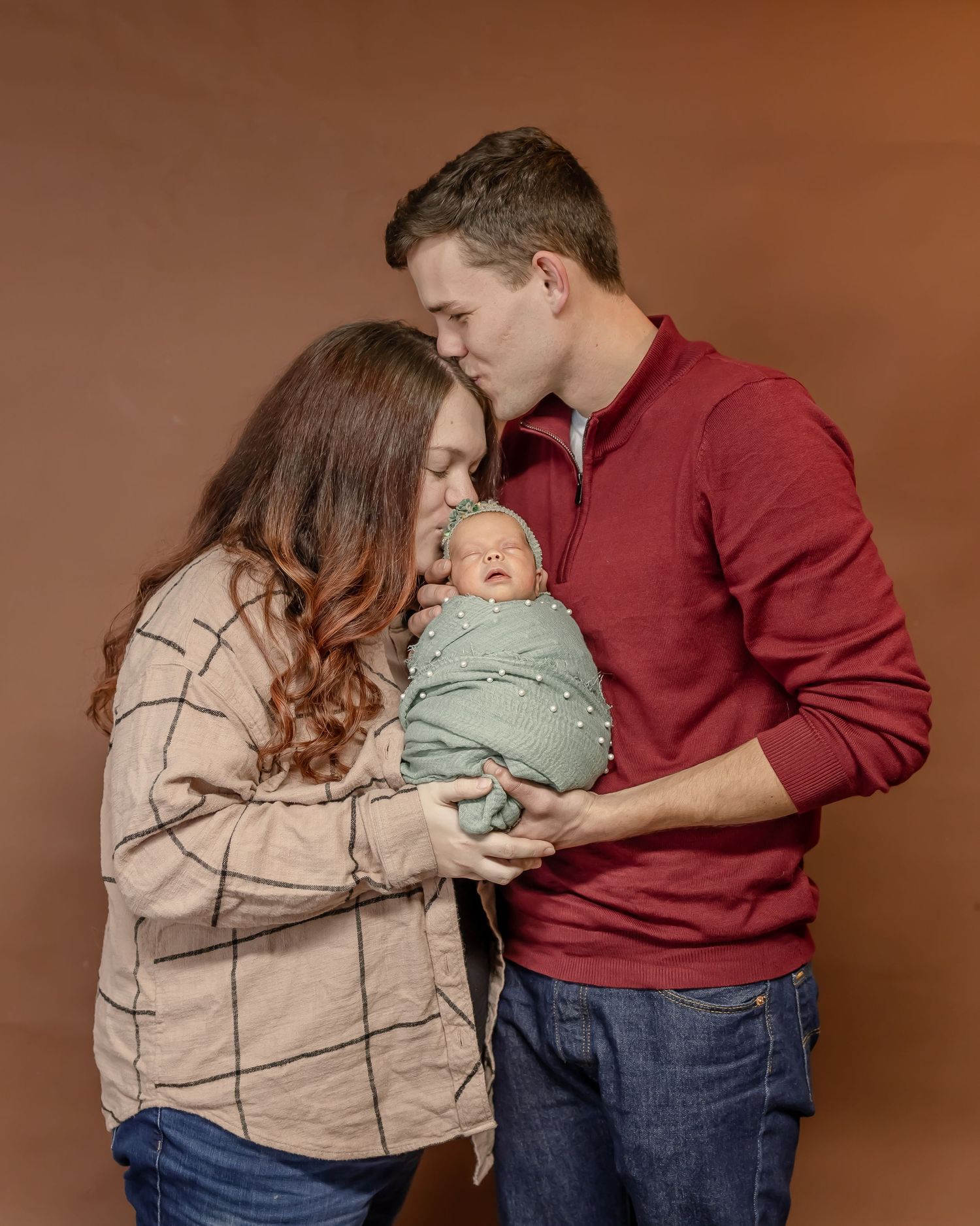 A tender family moment as parents snuggle with their newborn baby wrapped in a mint green blanket against a brown backdrop.