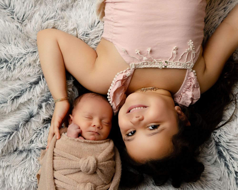 A peaceful newborn swaddled in beige fabric lies next to someone on a plush gray blanket.
