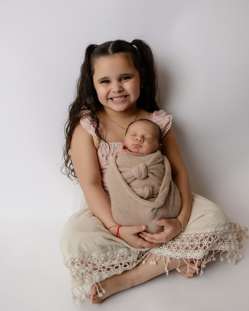 A young child in a lace-trimmed dress sits cross-legged while holding a swaddled newborn against a light background.