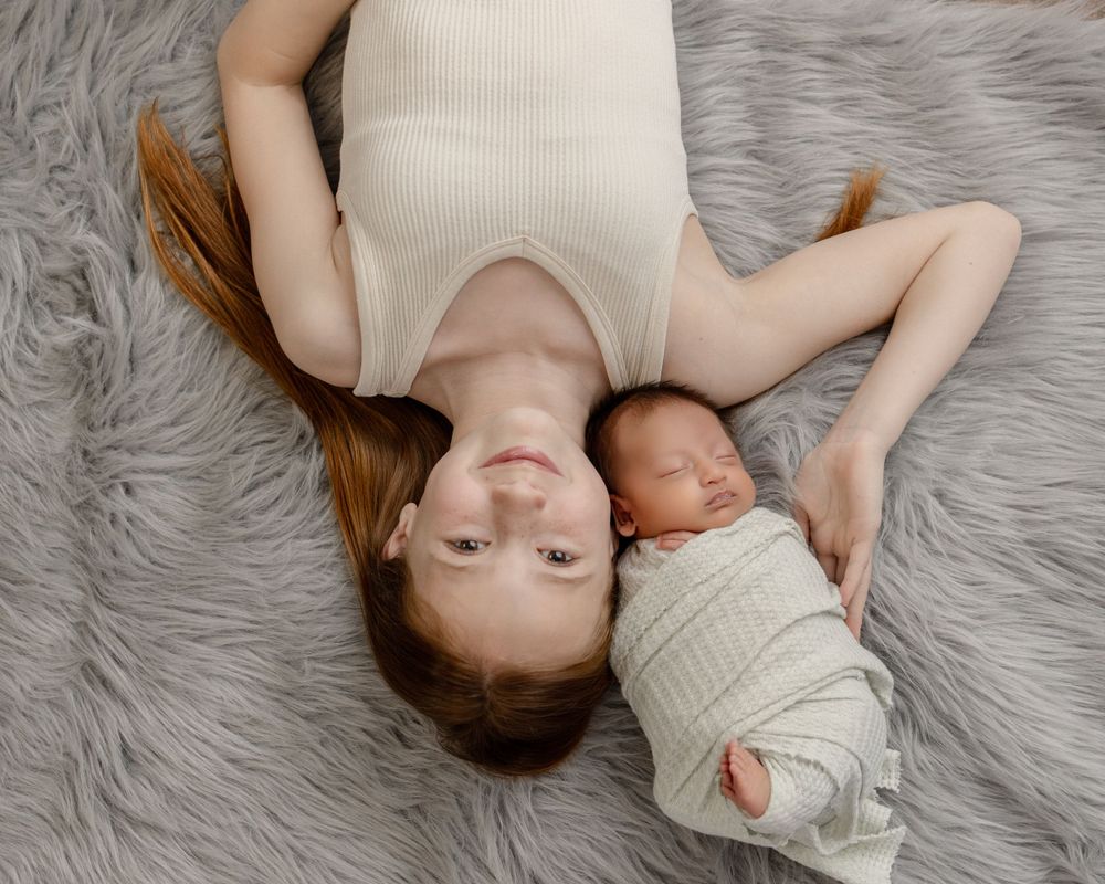 A person and infant wearing matching white knits lie together on a soft grey fur rug.