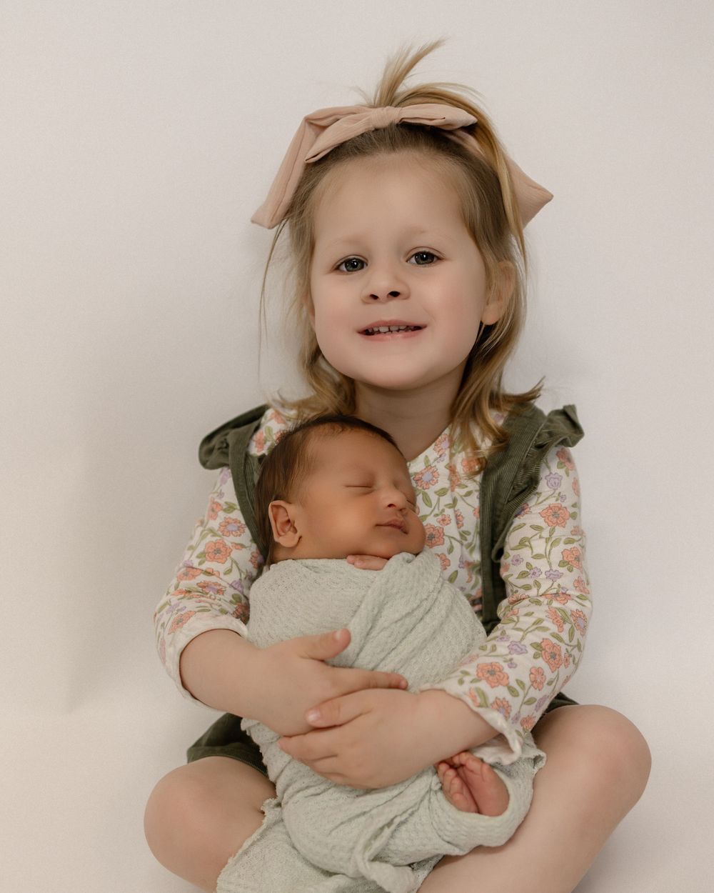 A tender sibling portrait shows gentle cradling of a sleeping infant on a neutral backdrop.