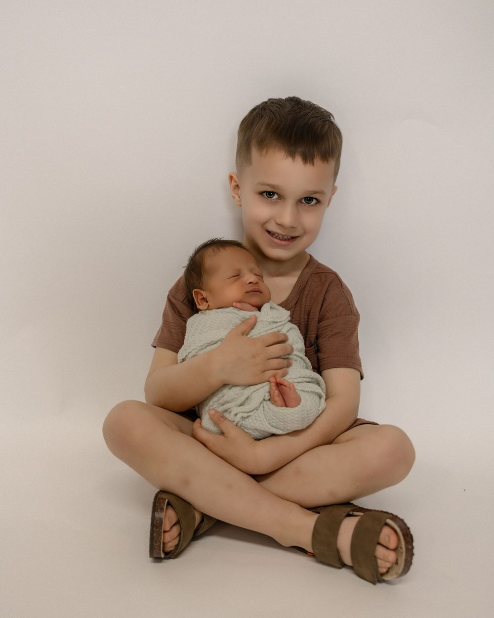 Two children sitting together on a light backdrop, one holding a small infant in a brown shirt and sandals.