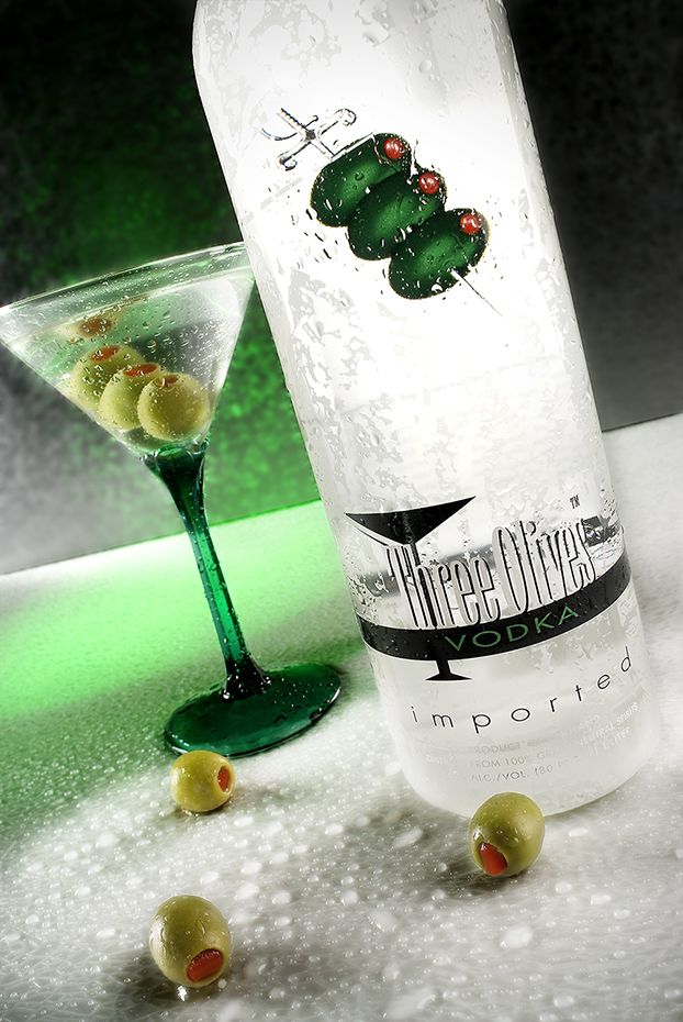 A martini glass and bottle of Three Olives vodka with green lighting and scattered olives on a frosty surface.