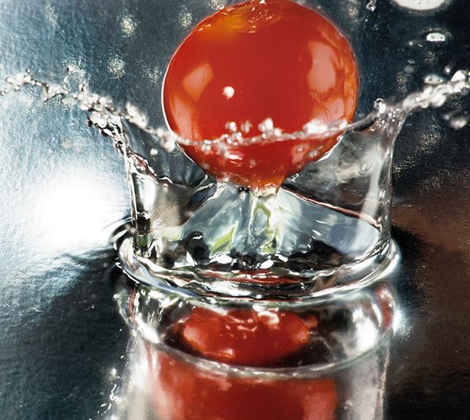 A bright red cherry splashing into clear liquid creating a dramatic crown-like water effect with reflections.
