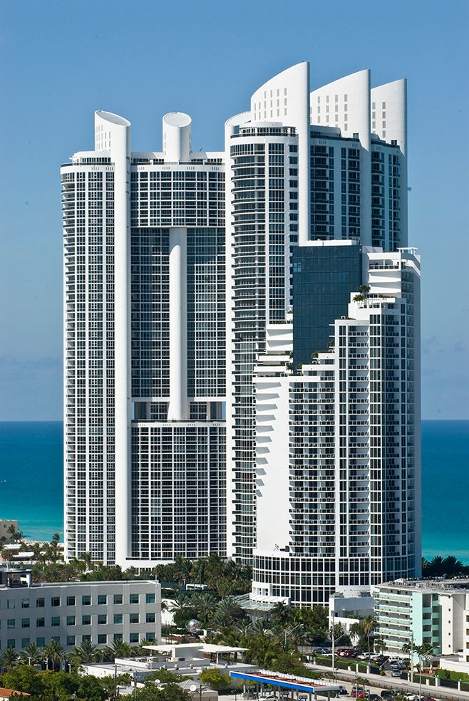 Modern white and blue oceanfront luxury condo tower rises above Miami Beach with dramatic architectural details and turquoise waters.