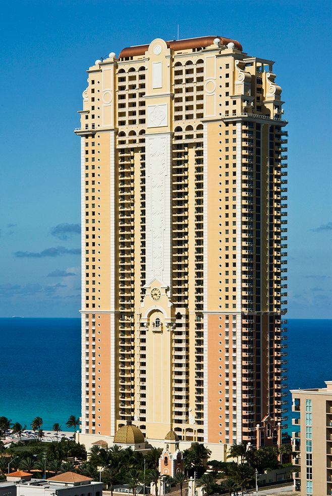 Luxury beachfront high-rise condominium tower with tan exterior stands tall against blue ocean and sky in sunny coastal setting.