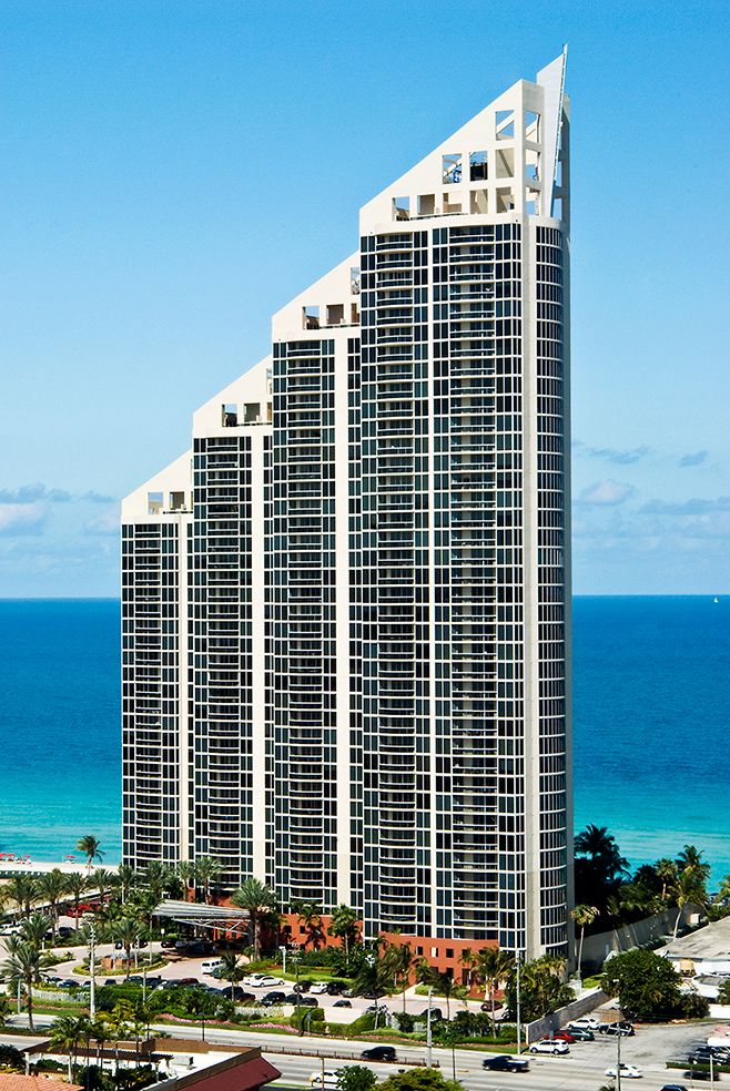 Luxury oceanfront condominium tower with slanted roof rises dramatically against blue sky and turquoise waters in Miami Beach.