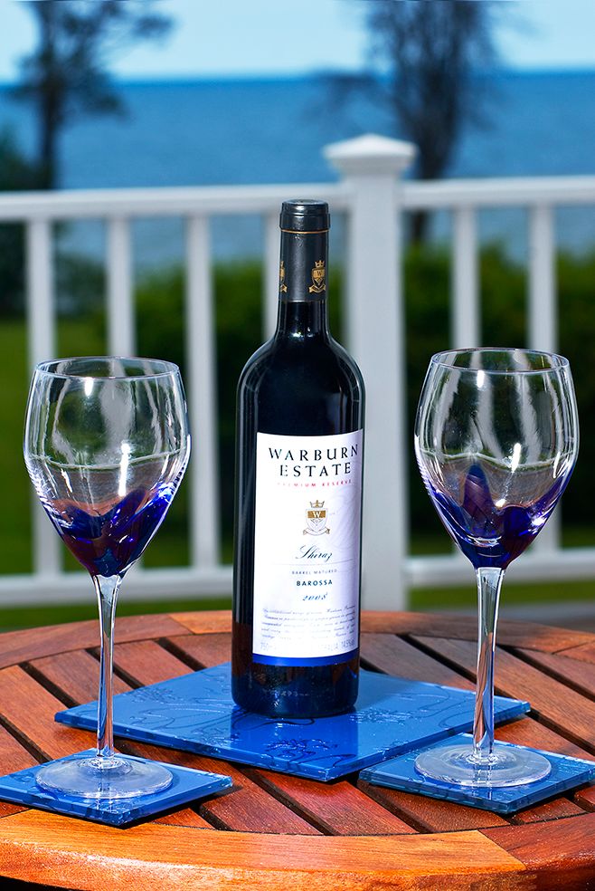 Bottle of Warburn Estate wine with two blue-tinted wine glasses on wooden table overlooking waterfront deck railing.