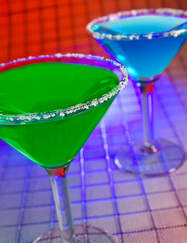 Two vibrant martini cocktails in glasses with sugar-rimmed edges, one bright green and one electric blue, on a grid surface.