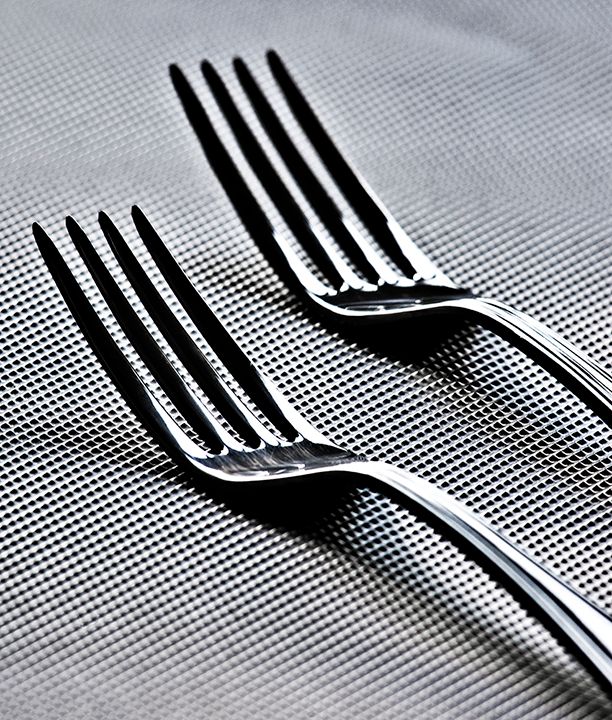 Two silver dinner forks arranged in parallel on a gray textured tablecloth with dramatic lighting and shadows.