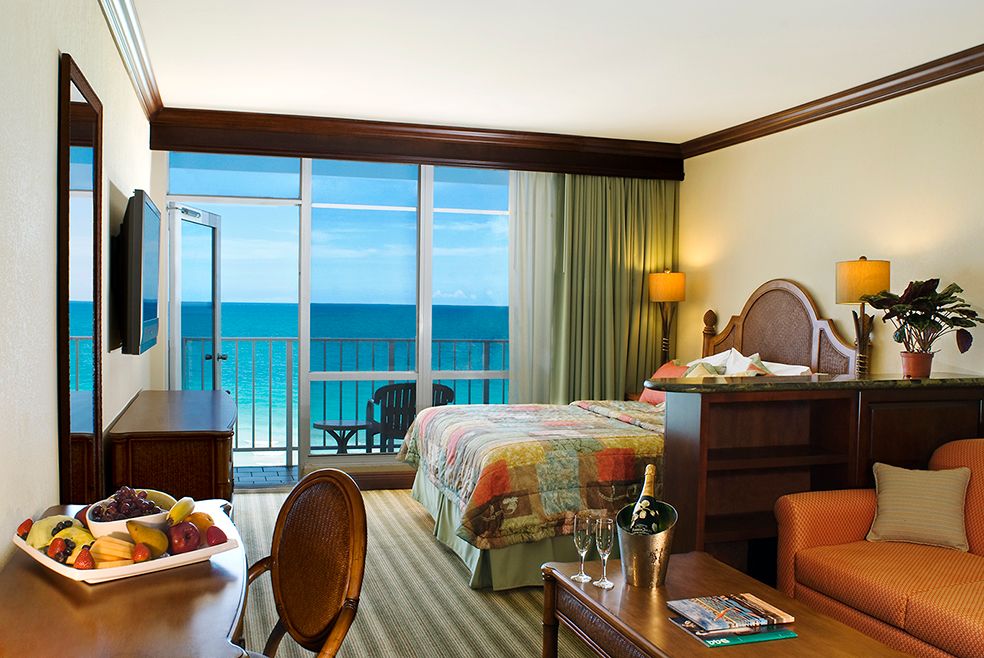 Luxurious oceanfront hotel room with king bed, private balcony, and stunning turquoise water views through floor-to-ceiling windows.