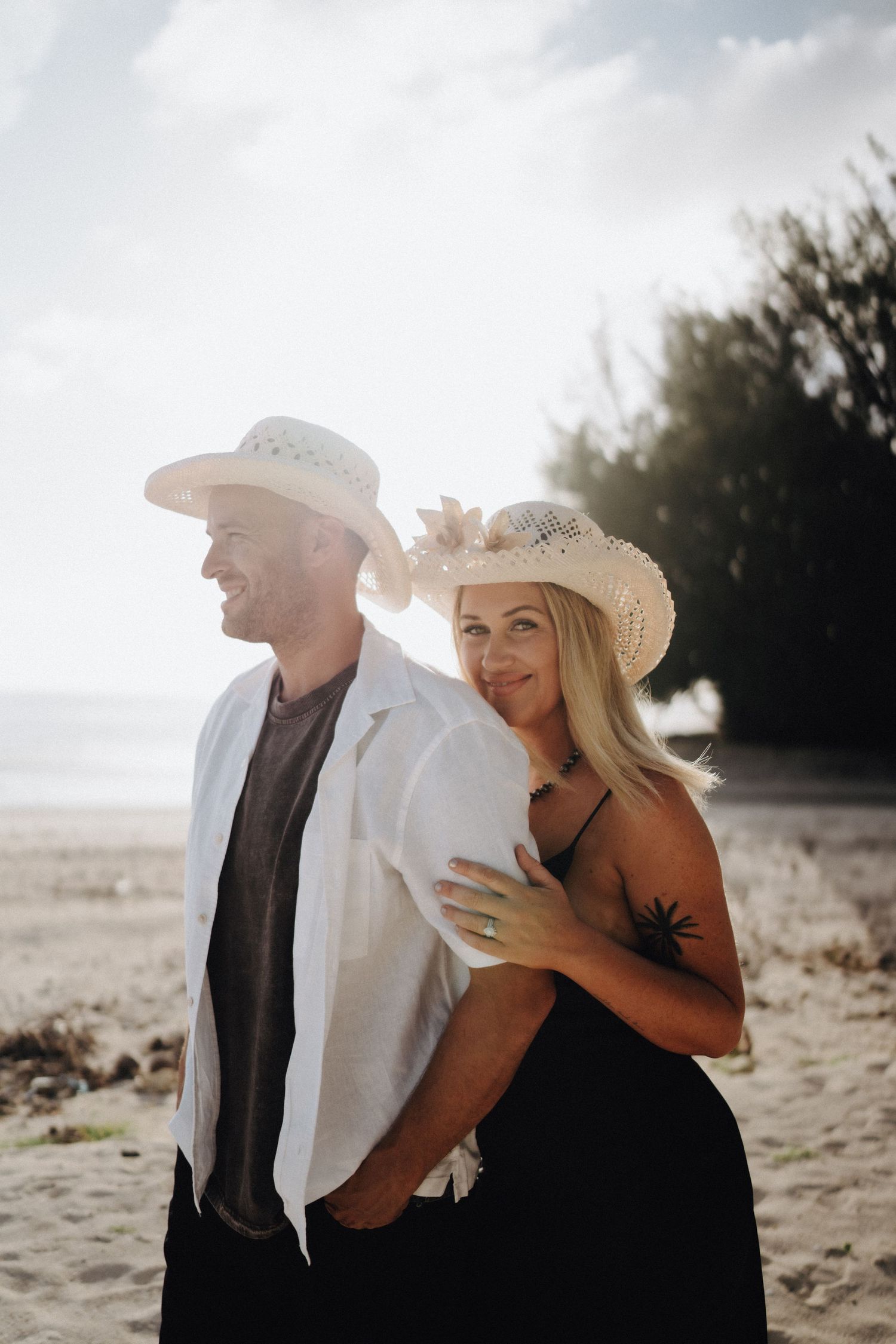 A couple wearing cowboy hats embrace on a scenic beach during a romantic photoshoot at sunset.