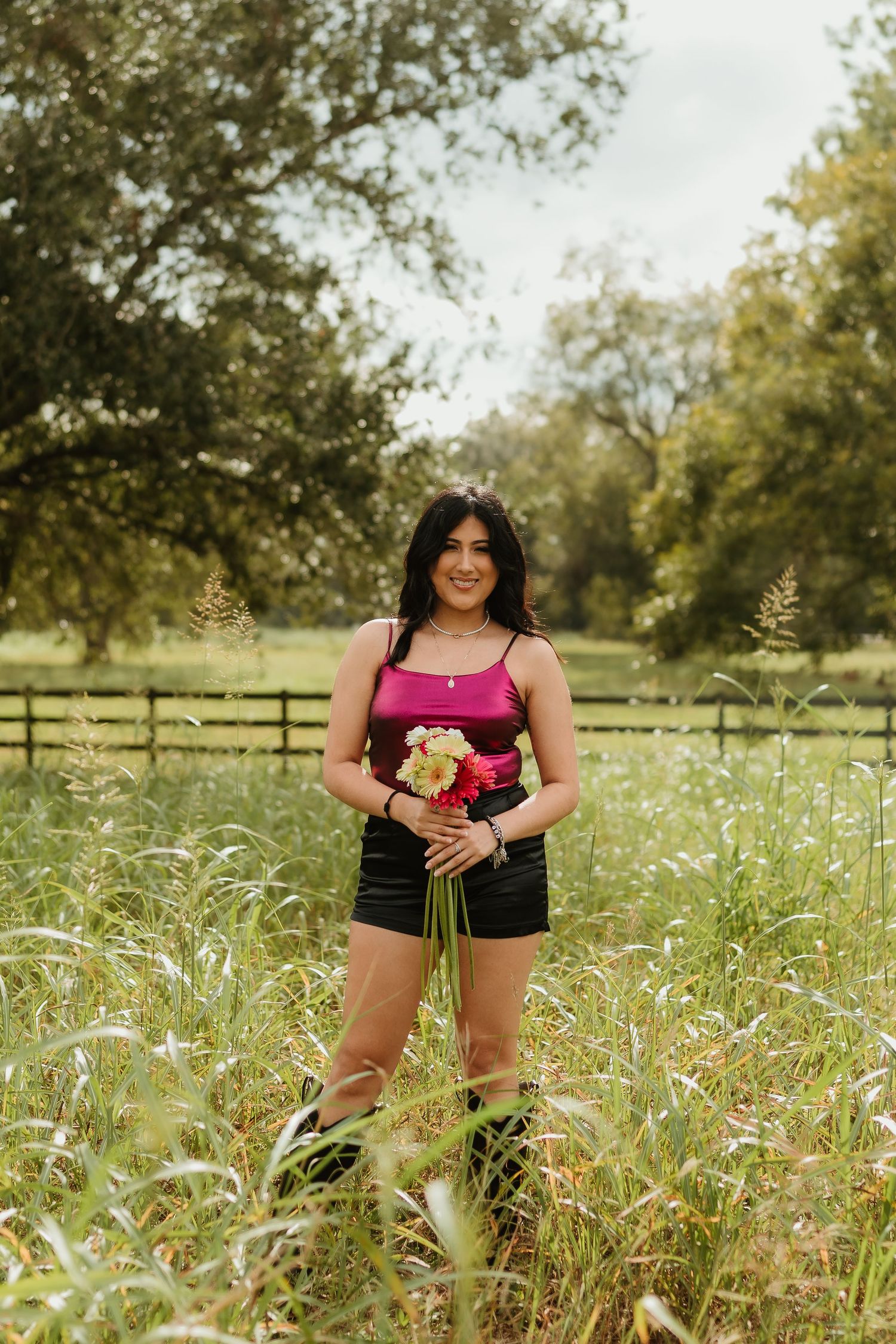 Woman in pink top poses with flowers in a sunny meadow with trees and fence in background.