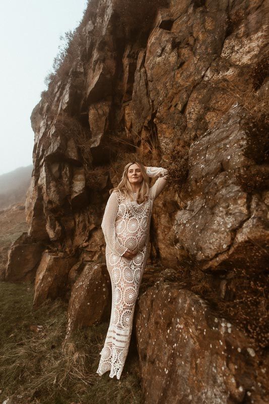 A person in a lace gown poses against a dramatic rocky cliff face during a misty outdoor photoshoot.