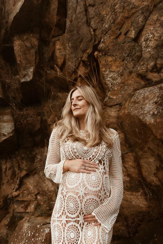 Pregnant woman in white lace dress poses against rocky cliff backdrop in warm tones.