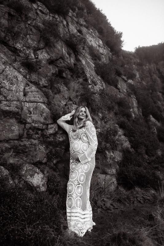 Black and white portrait of woman in lace dress standing against dramatic cliff face.