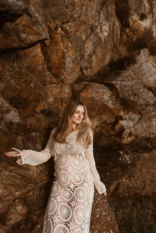 A woman in a white lace dress poses against a textured rock wall in warm earth tones, during a maternity photoshoot in Chorley.
