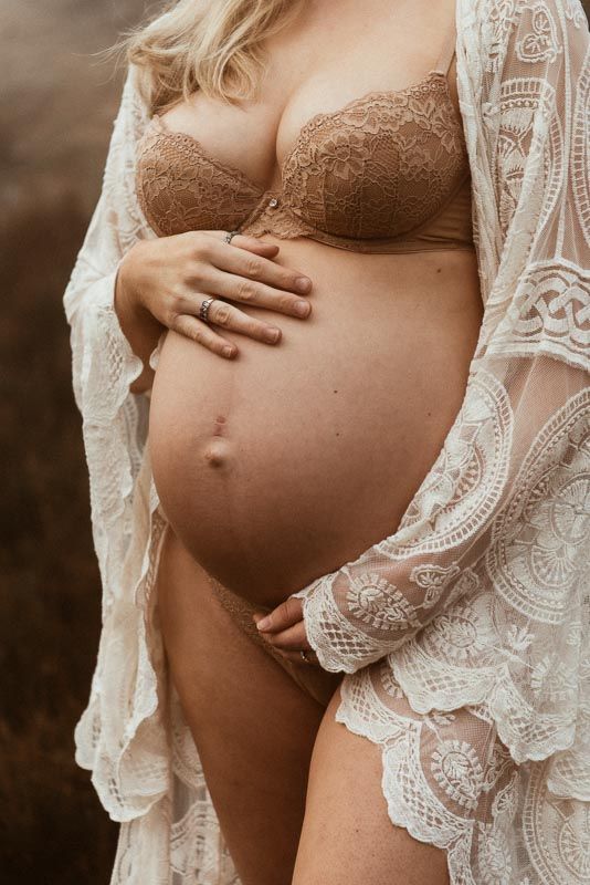 A pregnant woman in a brown lace bra and white sheer lace robe holds her bare belly in an intimate maternity photoshoot in Lancashire