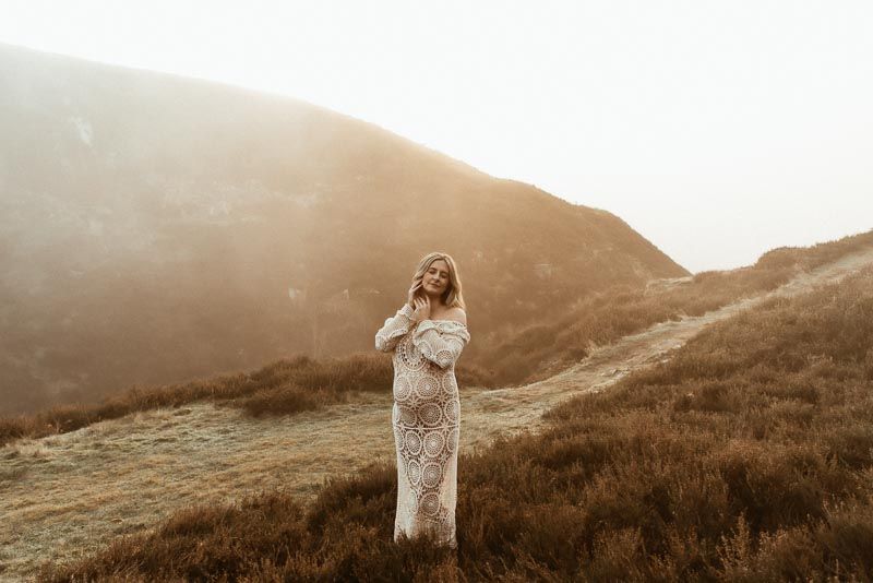 A woman in a lace gown stands on a misty mountainside during golden hour in a romantic outdoor setting.