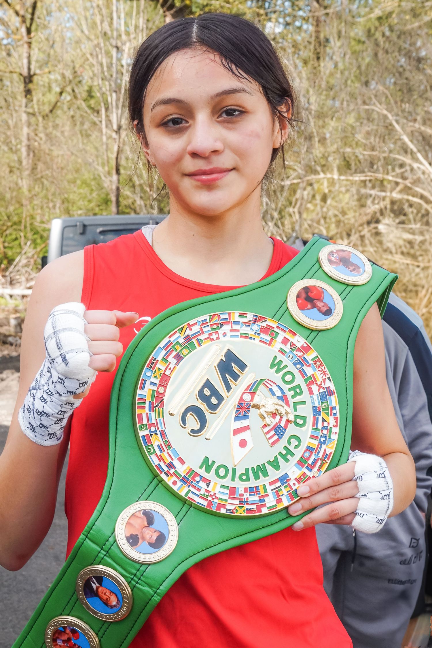 Young boxer poses with green WBC champion belt and white hand wraps in outdoor setting.