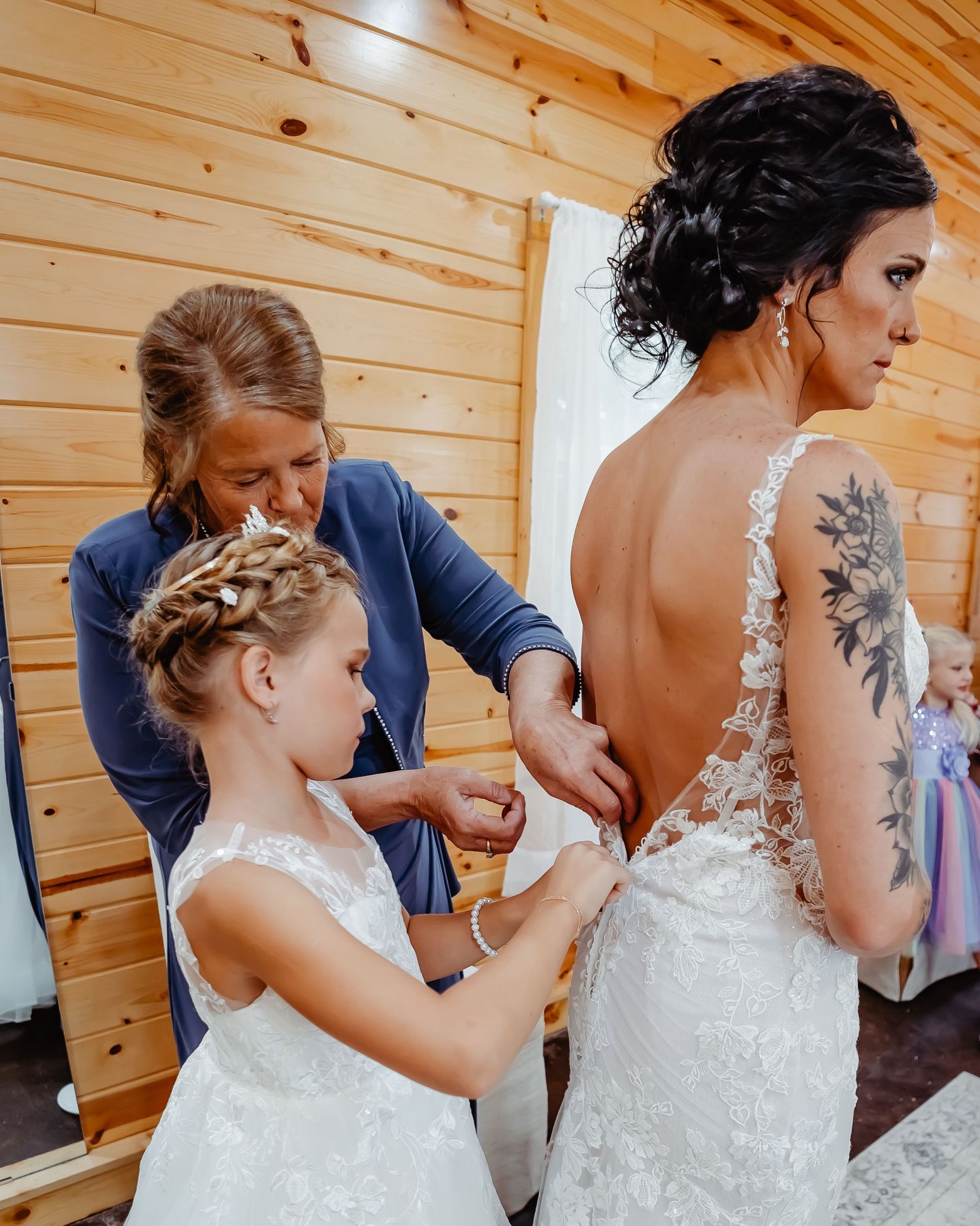 A touching wedding preparation moment shows a bride in lace dress being helped by others in a rustic wooden room.