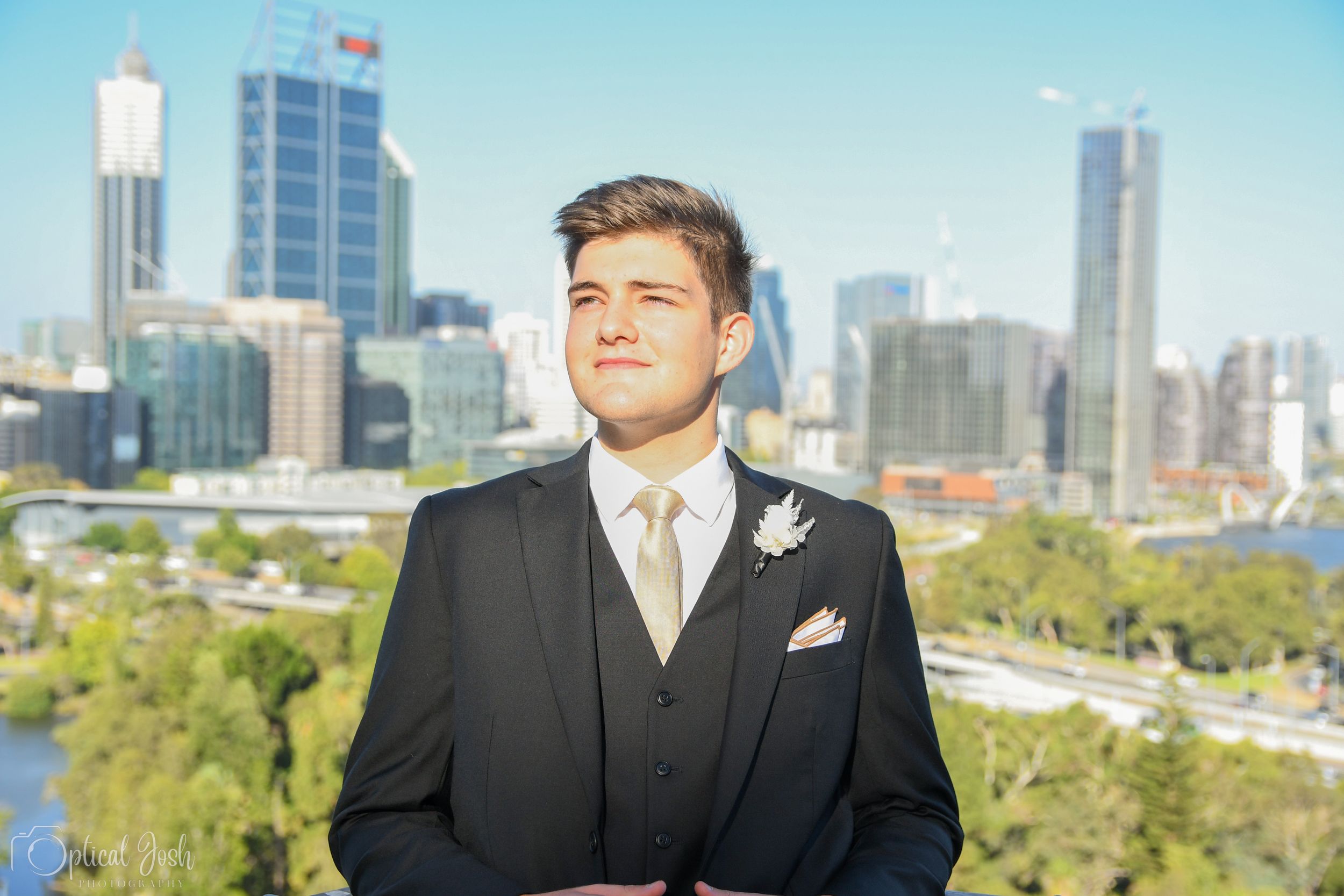 A well-dressed person in a formal suit stands against a city skyline backdrop on a sunny day.