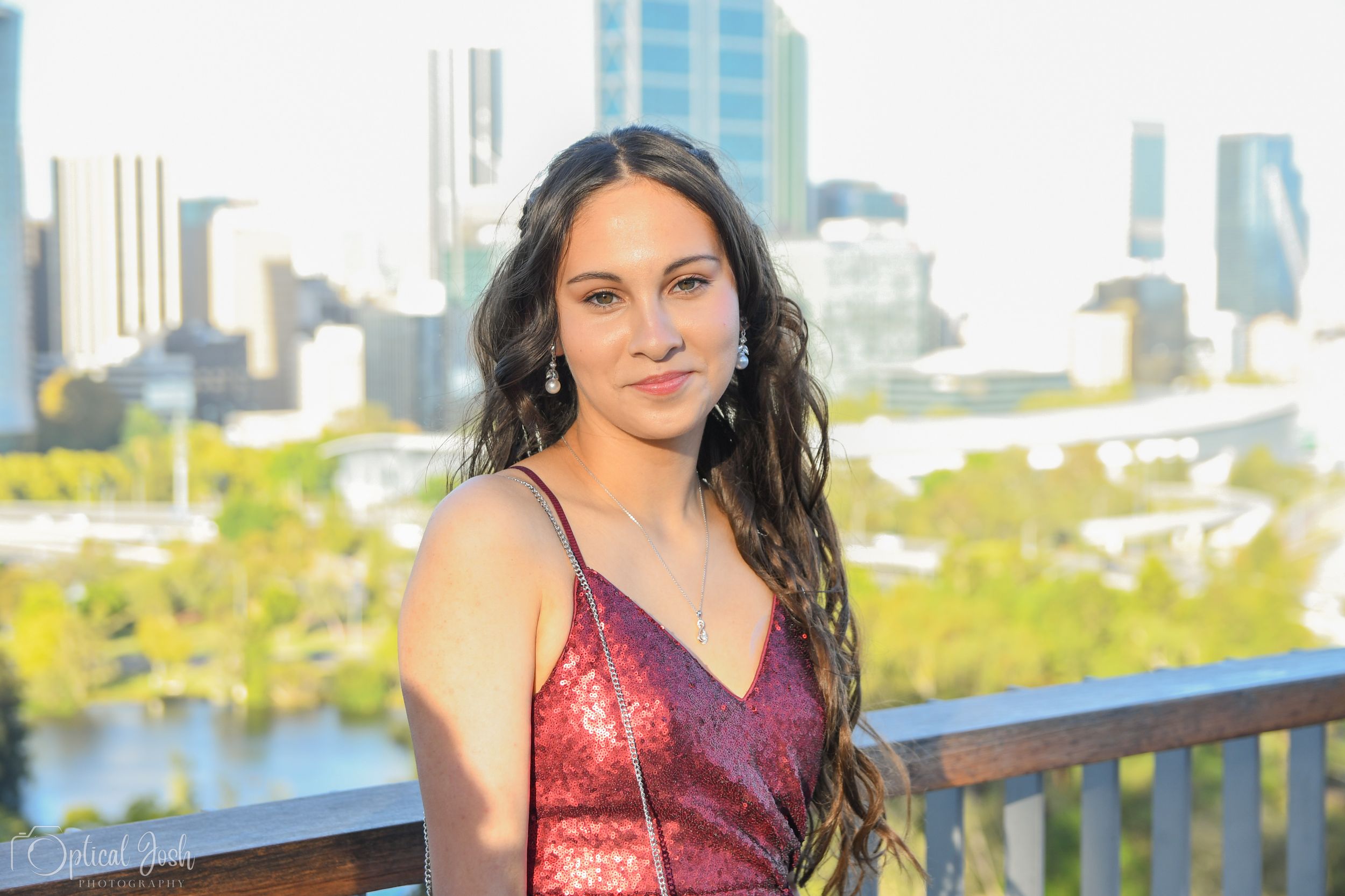 Woman in burgundy sequined dress poses on a balcony with a city skyline and green park in the background.