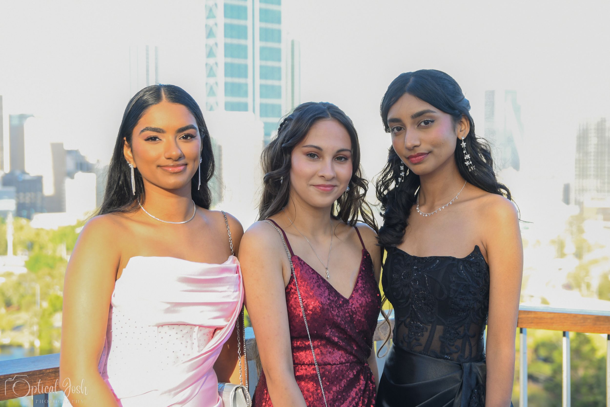 Three elegantly dressed friends pose together at a rooftop venue with city buildings visible in the background.