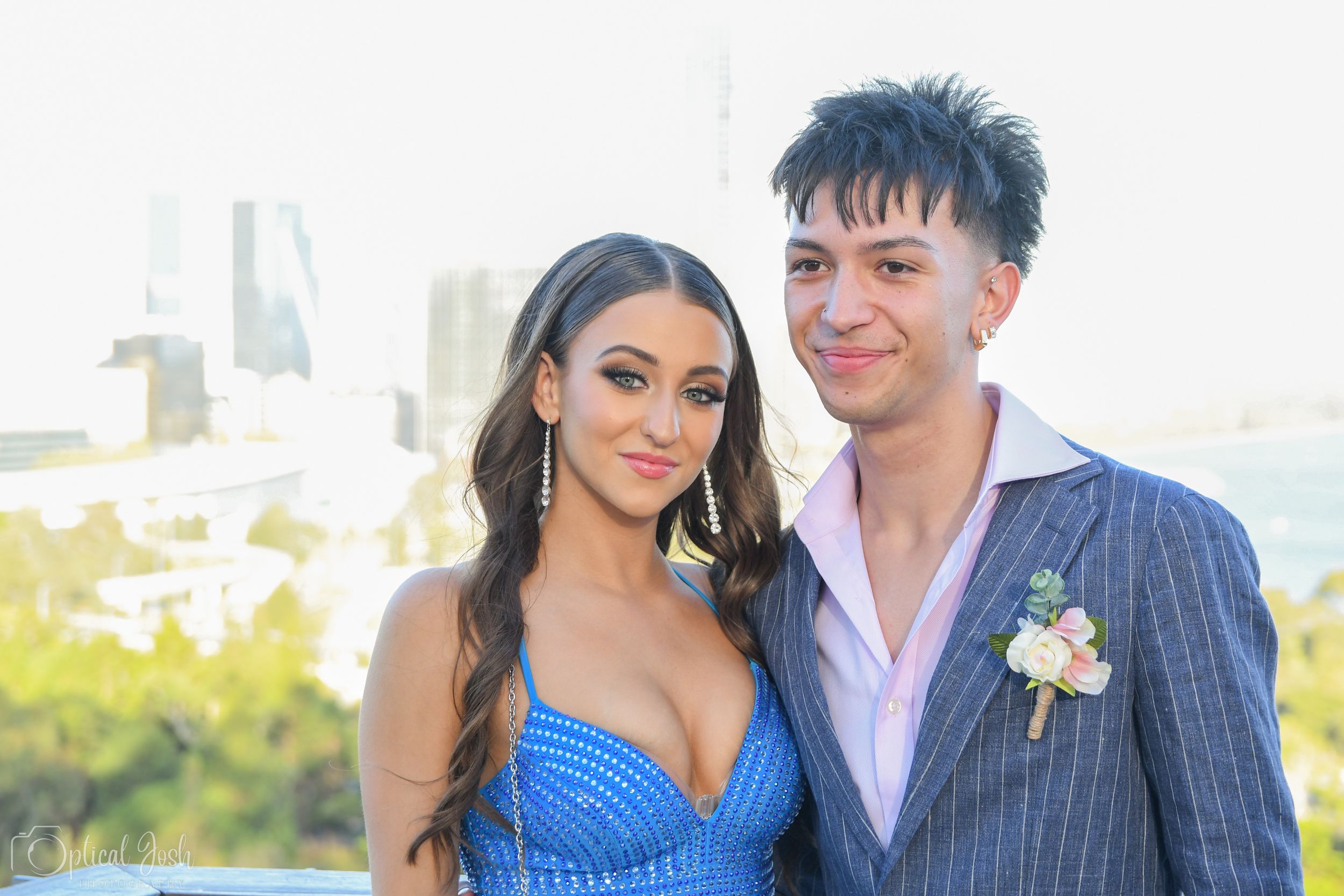 A young couple poses outdoors, with her wearing a blue dress and him in a blazer with boutonniere against a city backdrop.