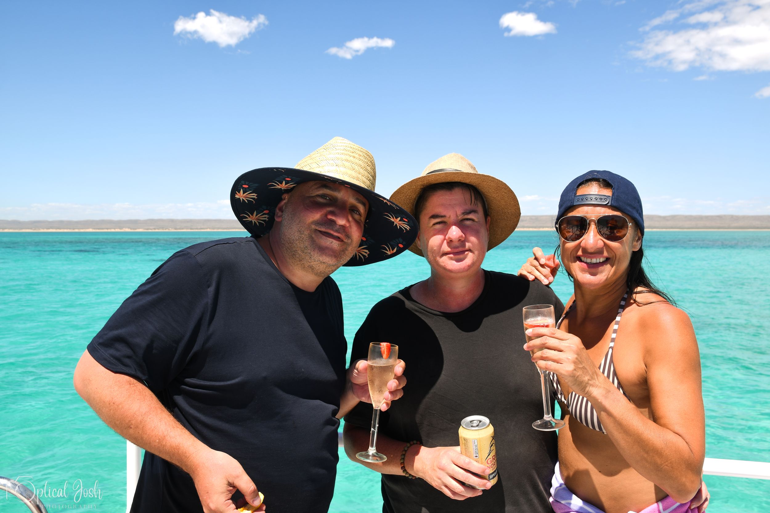Three people in hats enjoying drinks on a boat with bright turquoise tropical waters and blue sky in the background.