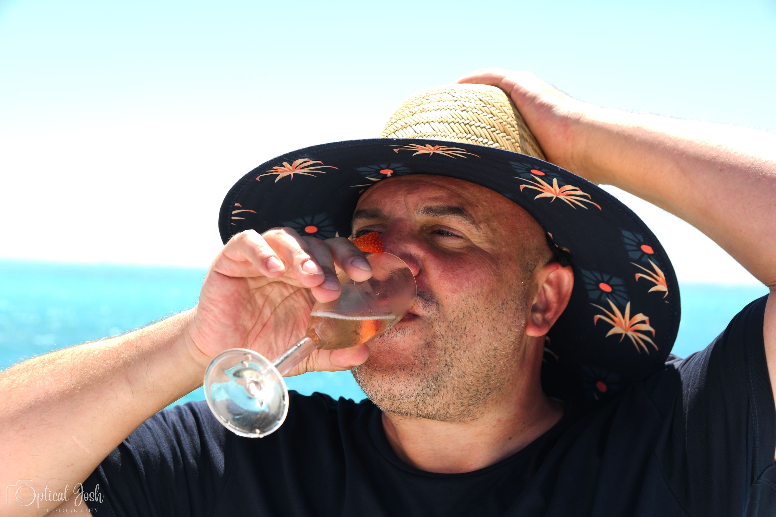 Someone in a palm tree print sun hat drinks from a wine glass against a bright blue ocean background.