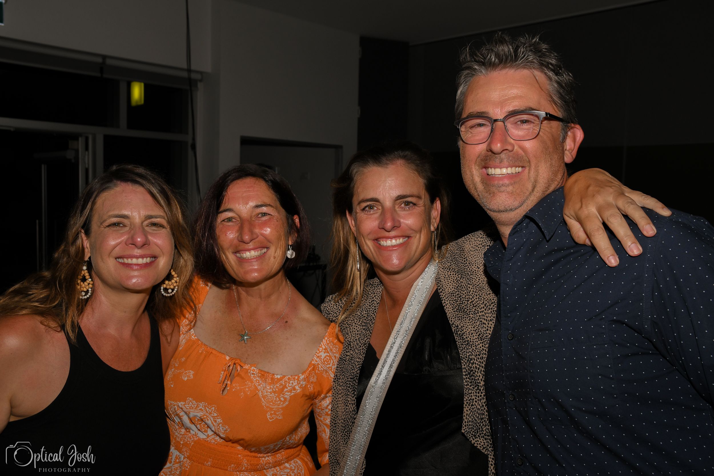 Four friends smiling and posing together at a dimly lit indoor evening gathering.