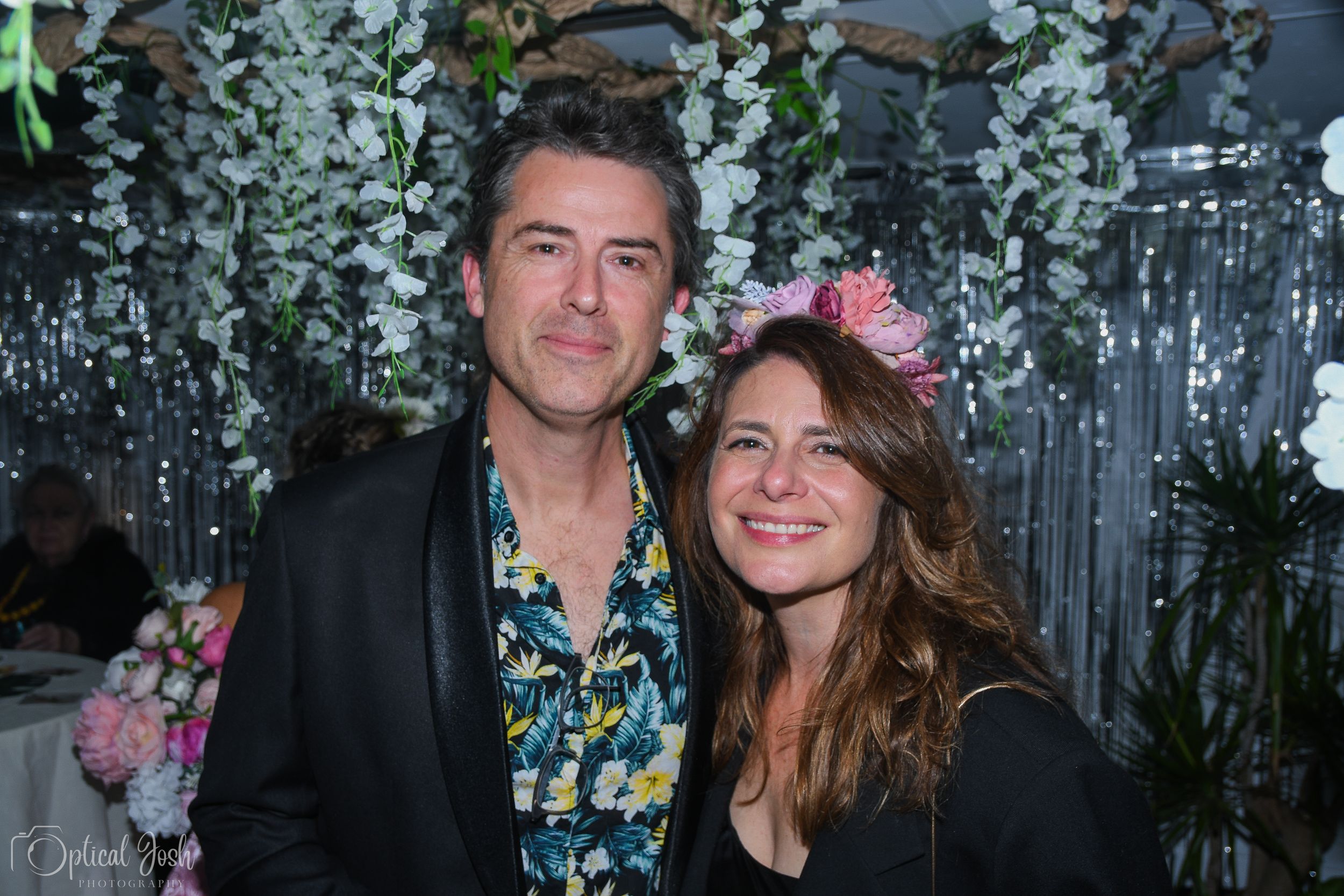 Two people smiling together at an event with decorative hanging floral arrangements in the background.