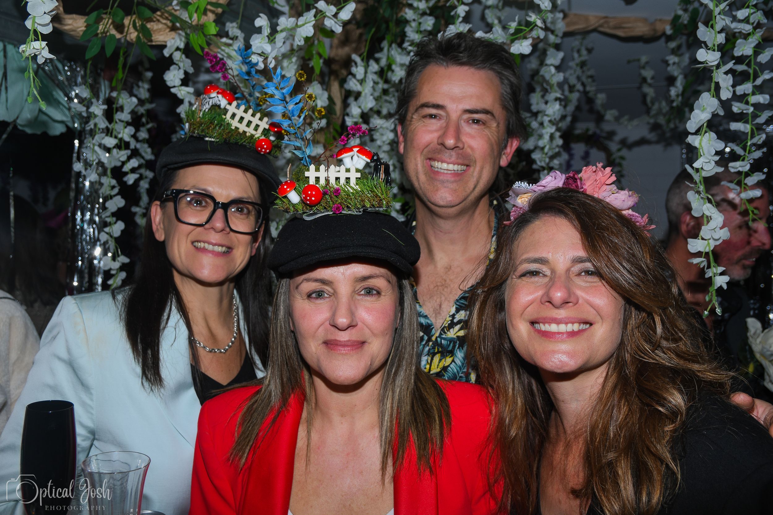 A group of friends smile together at a party with floral decorations and twinkling lights in the background.