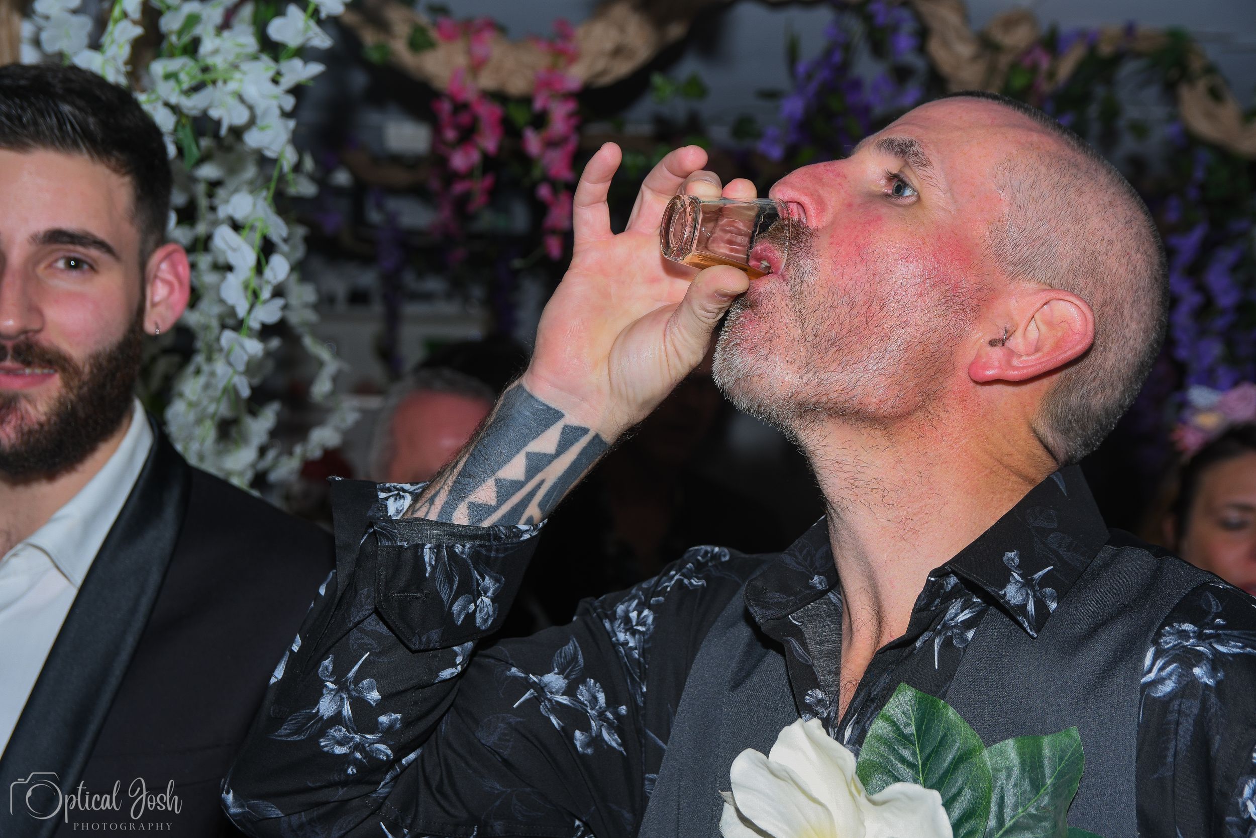 Man in dark floral shirt drinks from a shot glass at an event with decorative flowers in the background.